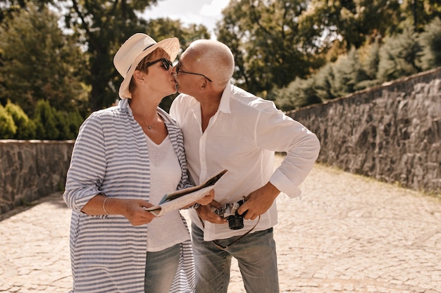 Beautiful woman with shirt blonde hair in hat and long striped blouse kissing with grey haired man in white long sleeve shirt with camera outdoor.