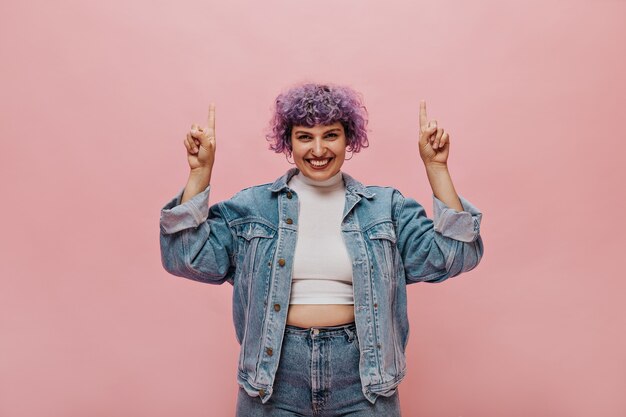 Beautiful woman with stylish haircut in white summer top and denim wide jacket is smiling. Wonderful lady in round earrings raises her fingers up.