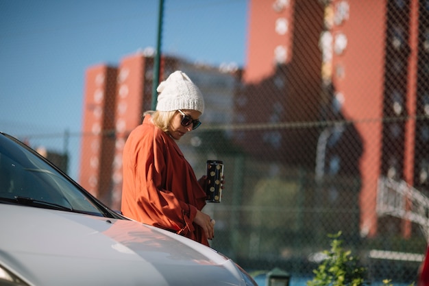 Beautiful woman with thermos leaning on car