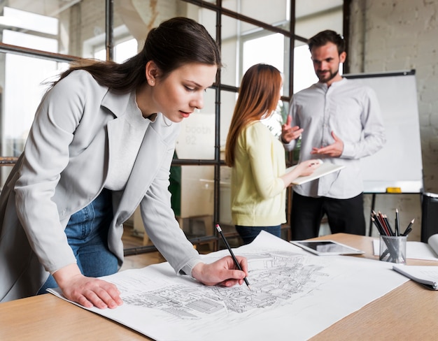 Beautiful woman working on blue print while her colleague discussing something on background