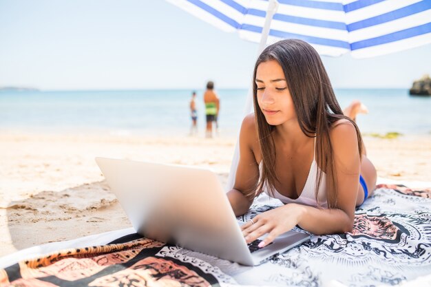 바다 근처 태양 우산 아래 해변에 누워있는 동안 노트북에서 온라인으로 작업하는 아름 다운 여자. 편안 하 고 프리랜서 인터넷 작업을 위해 노트북을 사용 하여 행복 하 게 웃는 프리랜서 소녀.