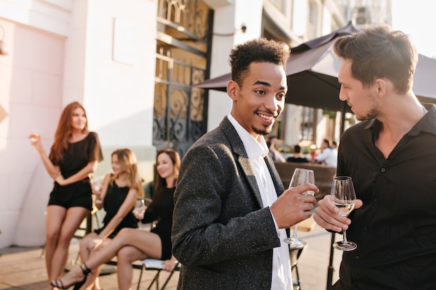 Beautiful women with long legs sitting at street cafe while guys on foreground clinking glasses