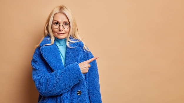 Beautiful wrinkled middle aged woman pointing at copy space on brown wall