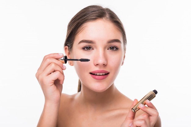 Beautiful young adult woman applying mascara on eyelashes isolated on white wall