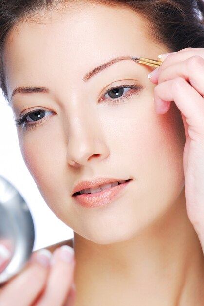 Beautiful young adult woman looking in mirror and plucking eyebrows