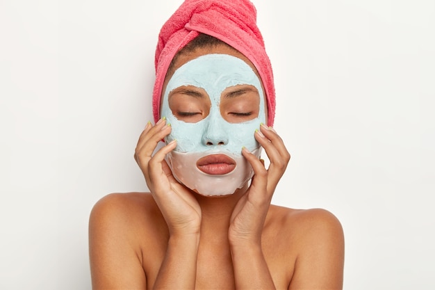 Beautiful young African American woman applies facial clay mask on face, touches skin gently, keeps eyes closed, wears wrapped towel on head, stands with bare shoulders, makes beauty treatments