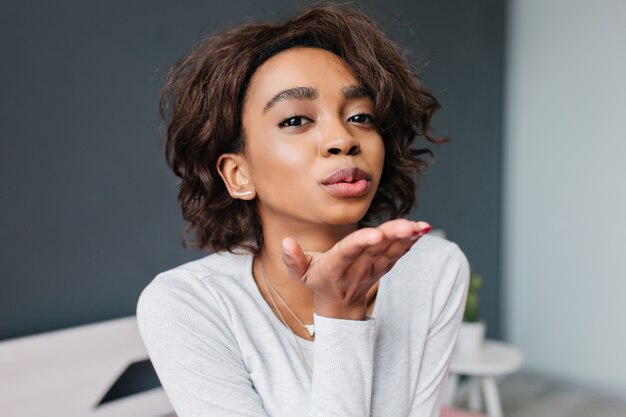 Beautiful young african girl with brown curly hair giving kiss in room with grey wall. Wearing light gray t-shirt with long sleeves, pendant triangle