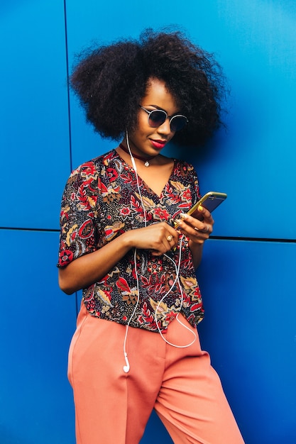 Beautiful young african woman in sunglasses and headphones, using a smartphone