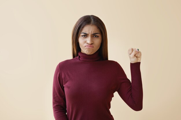 Beautiful young Afro American woman wearing turtle neck having determined angry expression, keeping fist pumped for feminine and independent strength, fighting against injustice and violence