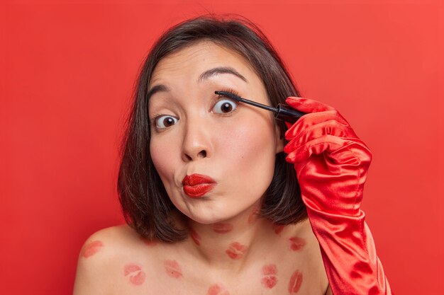 아름다운 젊은 아시아 여성이 속눈썹에 마스카라를 바르고 생생한 붉은 벽에 맨몸으로 데이트나 파티를 준비합니다.