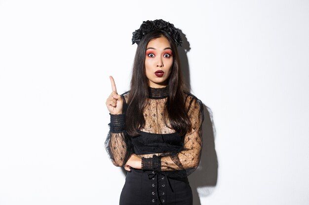 Beautiful young asian woman in black lace dress and gothic makeup have thought. Girl in witch costume raising finger, suggesting halloween idea.