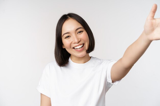 白い背景の上のTシャツに立っているselfieビデオチャットを取っているカメラ保持装置を見て笑っている美しい若いアジアの女性