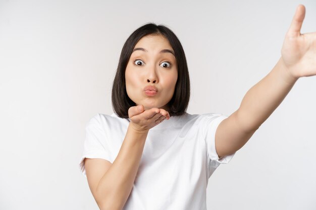 Beautiful young asian woman smiling looking at camera holding device taking selfie video chat standing in tshirt over white background