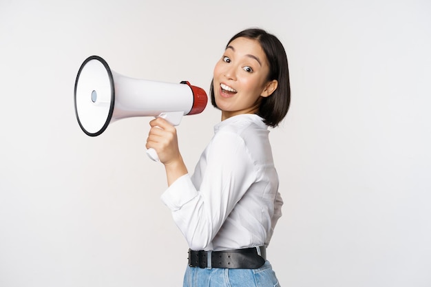 Beautiful young asian woman talking in megaphone screams in speakerphone and smiling making announcement shout out information standing over white background