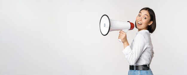 Beautiful young asian woman talking in megaphone screams in speakerphone and smiling making announcement shout out information standing over white background