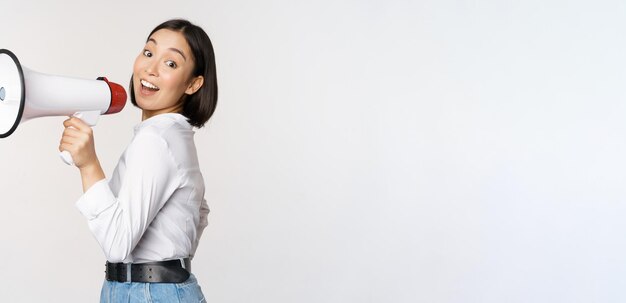 Beautiful young asian woman talking in megaphone screams in speakerphone and smiling making announcement shout out information standing over white background