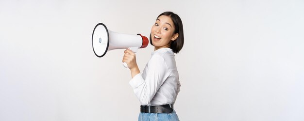 Beautiful young asian woman talking in megaphone screams in speakerphone and smiling making announcement shout out information standing over white background