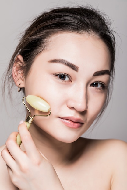 Beautiful young asian woman using a jade face roller on her flawless skin. Beauty face closeup. Conceptual of facial treatments with semi precious stones. isolated on gray with copy space