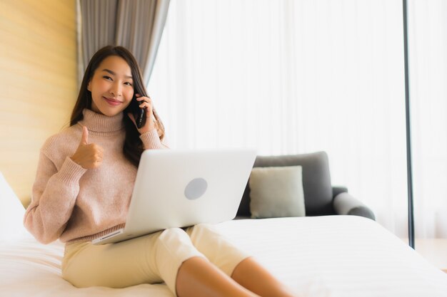 침대에서 휴대 전화로 노트북을 사용하는 아름 다운 젊은 아시아 여자