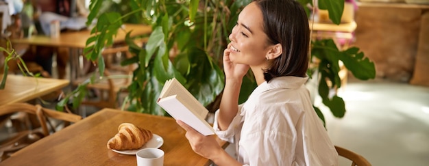 무료 사진 손에 책을 들고 카페에 앉아 커피를 마시고 먹는 아름다운 젊은 아시아 여성