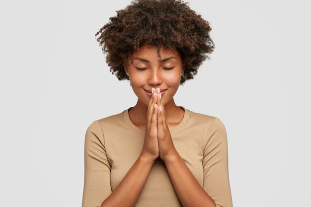 Beautiful young black woman stands in meditative pose, enjoys peaceful atmosphere, holds hands in praying gesture