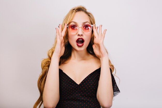 Beautiful young blonde girl with surprised face, young woman with opened mouth. Wearing pink cool glasses, black dress, has nice curly hair.