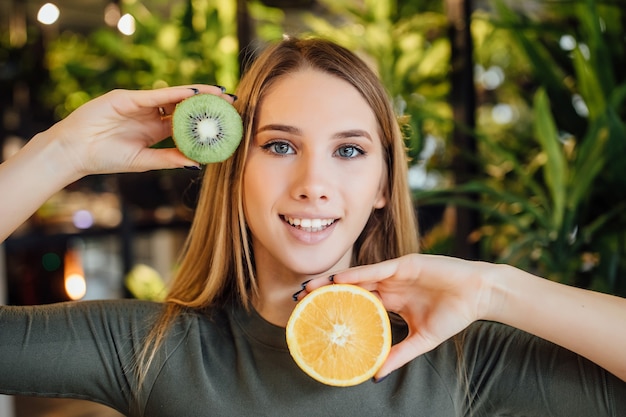 Beautiful young blonde woman holding piece of avocado and kiwi in front of her eye and touching her face