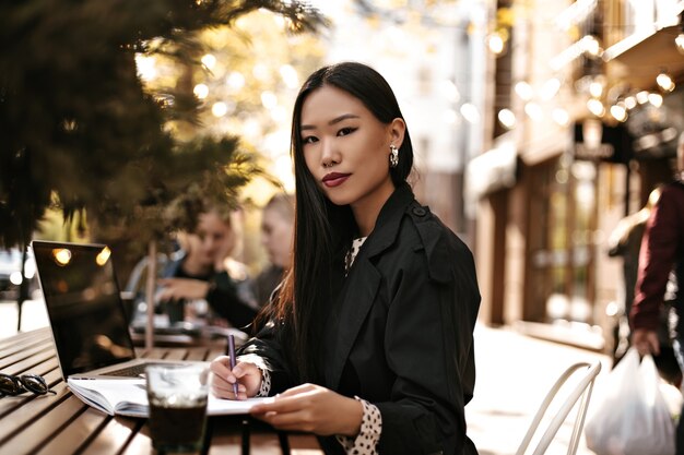 Beautiful young brunette Asian woman in black trench coat looks into camera, sits at wooden desk outside and makes notes in notebook