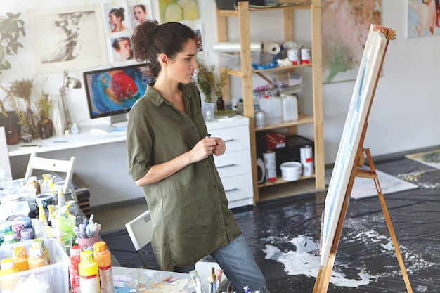 Beautiful young brunette female painter dressed casually standing in front of her painting, studying her picture with appraising look, thinking of what colors to add. Art and creativity concept