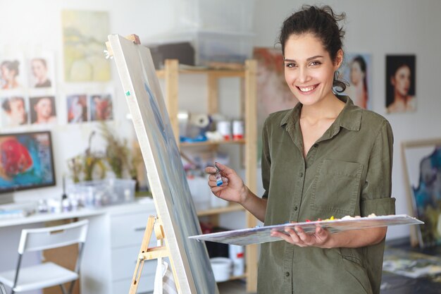 Beautiful young brunette female painter dressed casually while working in her workshop, standing near easel, creating picture with colorful watercolors