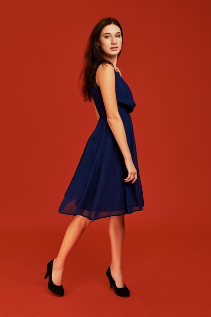 Beautiful young brunette woman in elegant blue cocktail dress and black high heels is posing for the camera. Side view. Full length studio shot on red background
