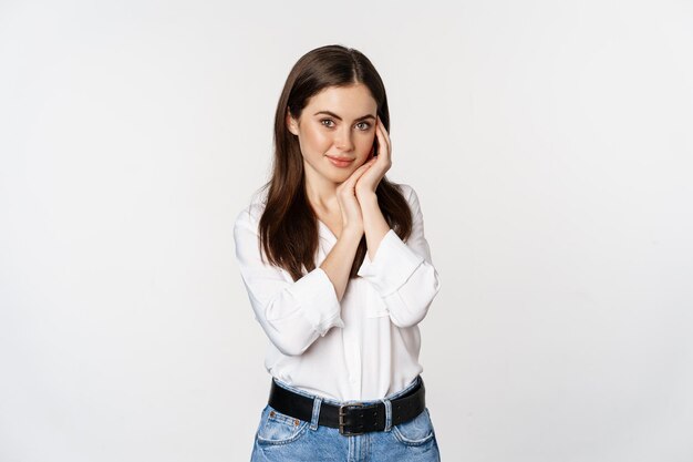 Beautiful young brunette woman, gently touches her face skin and smiling coquettish, tender pose against studio white background