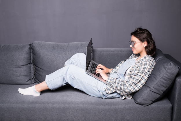 Красивая молодая брюнетка женщина дома, сидя на диване или диване, используя свой портативный компьютер и улыбаясь