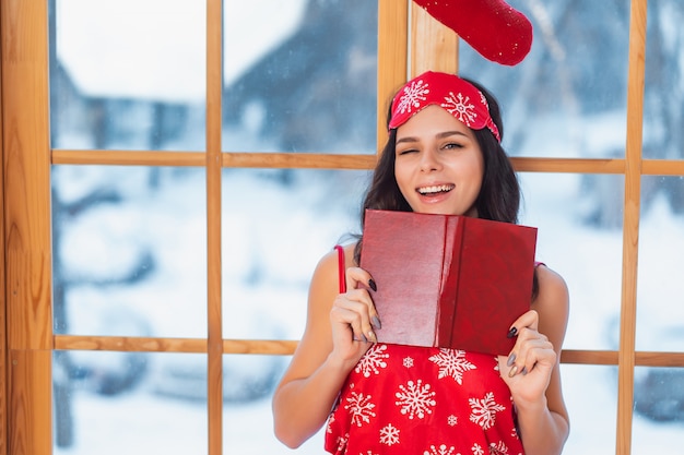 Free photo beautiful young brunette woman wearing red pajamas and reading by the window