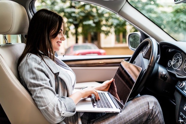 Beautiful young business woman using laptop and phone in the car.