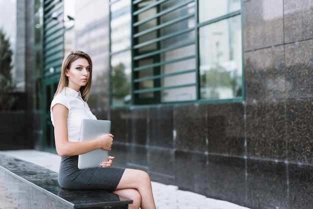 Beautiful young businesswoman holding laptop sitting on bench at outdoors