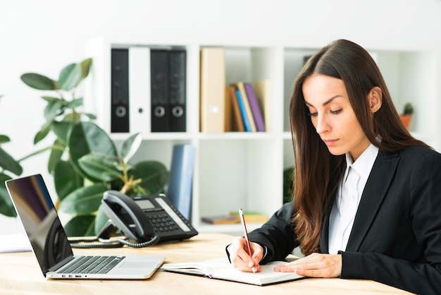 Beautiful young businesswoman writing with pencil on diary with laptop over desk