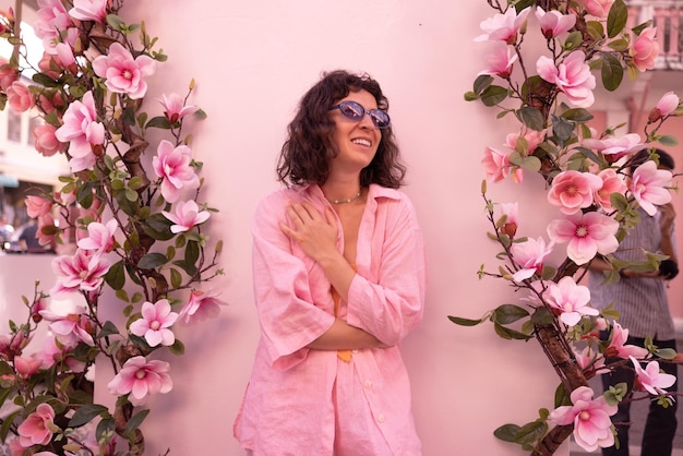 Beautiful young caucasian brunette woman in glasses and pink shirt is smiling standing on pink background with flowers