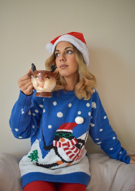 Beautiful young Caucasian female in cute blue Christmas outfit and Santa hat drinking hot chocolate