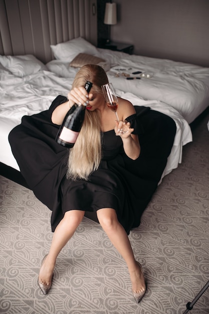 Foto gratuita bella giovane donna caucasica con lunghi capelli biondi, bel viso, trucco luminoso orecchini luminosi in abito nero lungo si siede sul grande letto bianco e beve vino