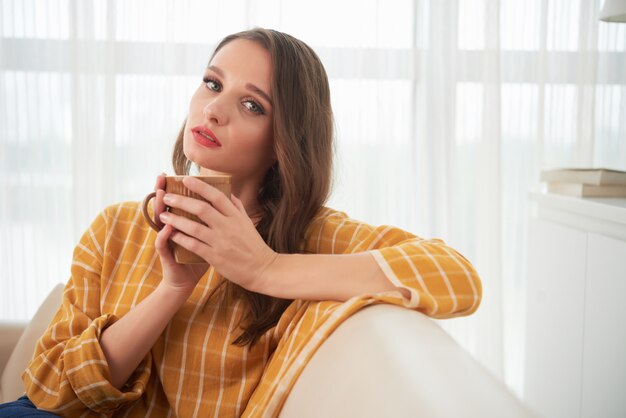 お茶を一杯で自宅のソファでポーズ美しい若い白人女性