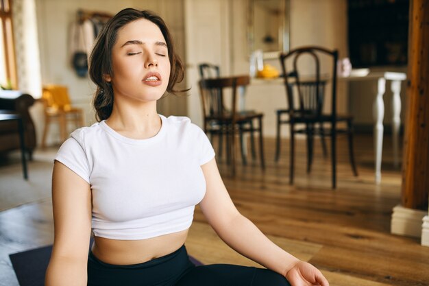 Beautiful young Caucasian woman with muscular curvy body sitting in lotus posture at home keeping eyes closed, meditating during yoga practive, doing body scanning, concentrating on breathing