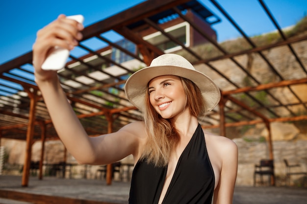 Beautiful young cheerful girl in hat rests at morning beach and makes selfie