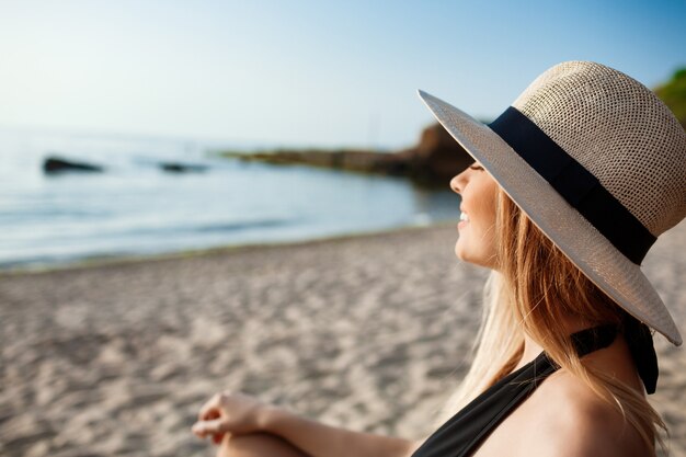 Красивая молодая жизнерадостная девушка в шляпе лежит на утреннем пляже