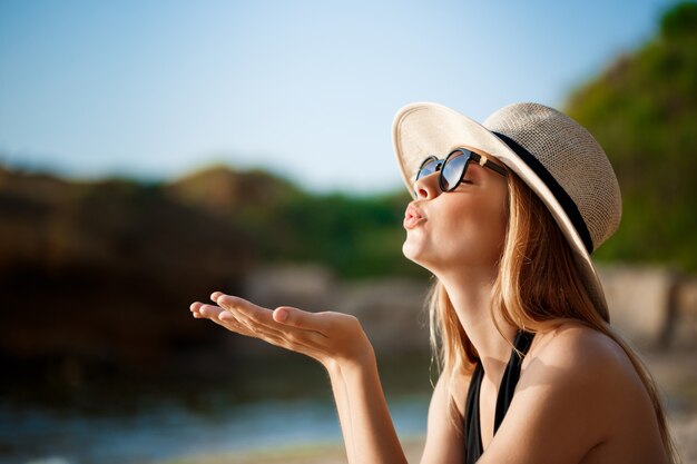 メガネと帽子を身に着けている美しい陽気な少女は朝のビーチにかかっています。
