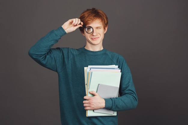 Beautiful young cheerful male student in stylish green sweater holding magnifier in front of eye and lot of notebooks, with happy and relaxed expression. Black wall