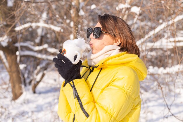 Beautiful young cheerful woman in a snowy landscape winter forest in sunglasses with a cup filled with snow having fun