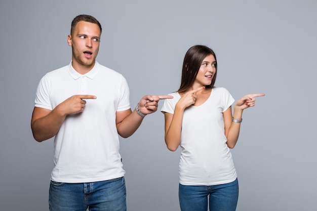 何かを示す白いTシャツに身を包んだ明るい灰色の背景に分離されたカジュアルな服装の美しい若いカップル