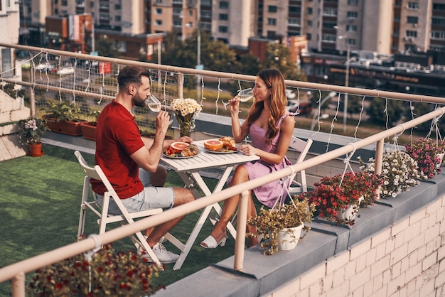 Красивая молодая пара в повседневной одежде наслаждается романтическим ужином и пьет шампанское, сидя во внутреннем дворике на крыше на открытом воздухе Premium Фотографии