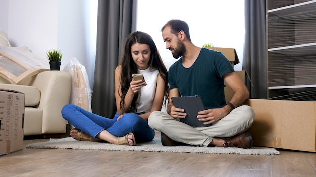 Beautiful young couple just moved in new flat. They are sitting on the floor and searching online for furniture